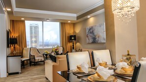 Une chambre d’hôtel comprenant un coin repas, un canapé, une table dressée pour deux personnes, un lustre et une vue sur la ville à travers de grandes fenêtres.