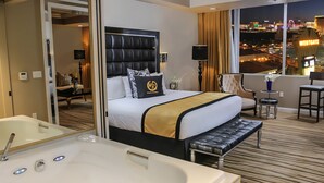 Une chambre d’hôtel avec un grand lit, un coin salon avec un fauteuil et une table, une baignoire et une vue sur la ville la nuit.