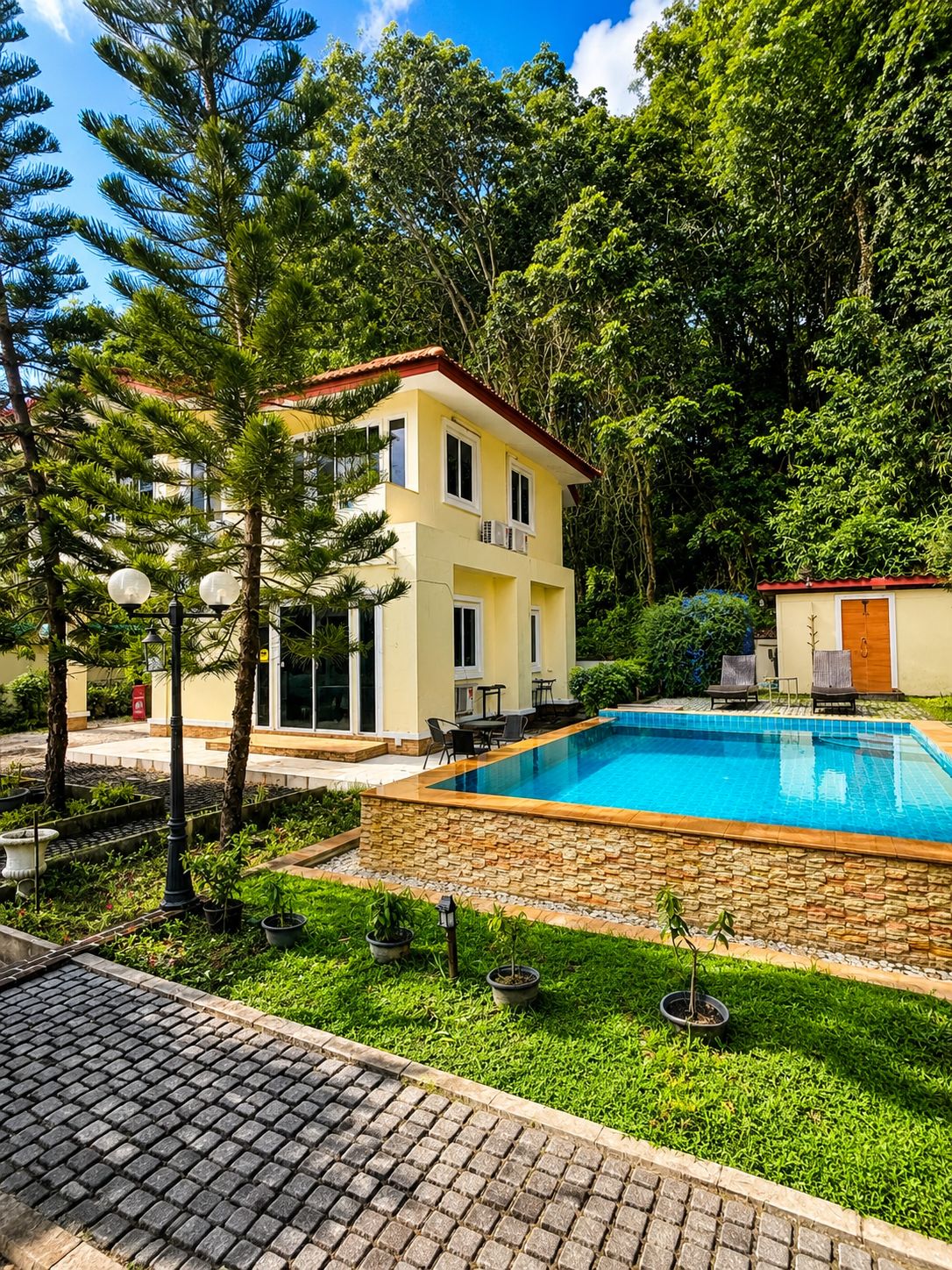 A two-story building with a swimming pool, surrounded by greenery and a paved area.