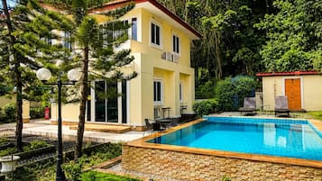 A two-story building with a swimming pool, surrounded by greenery and a paved area.