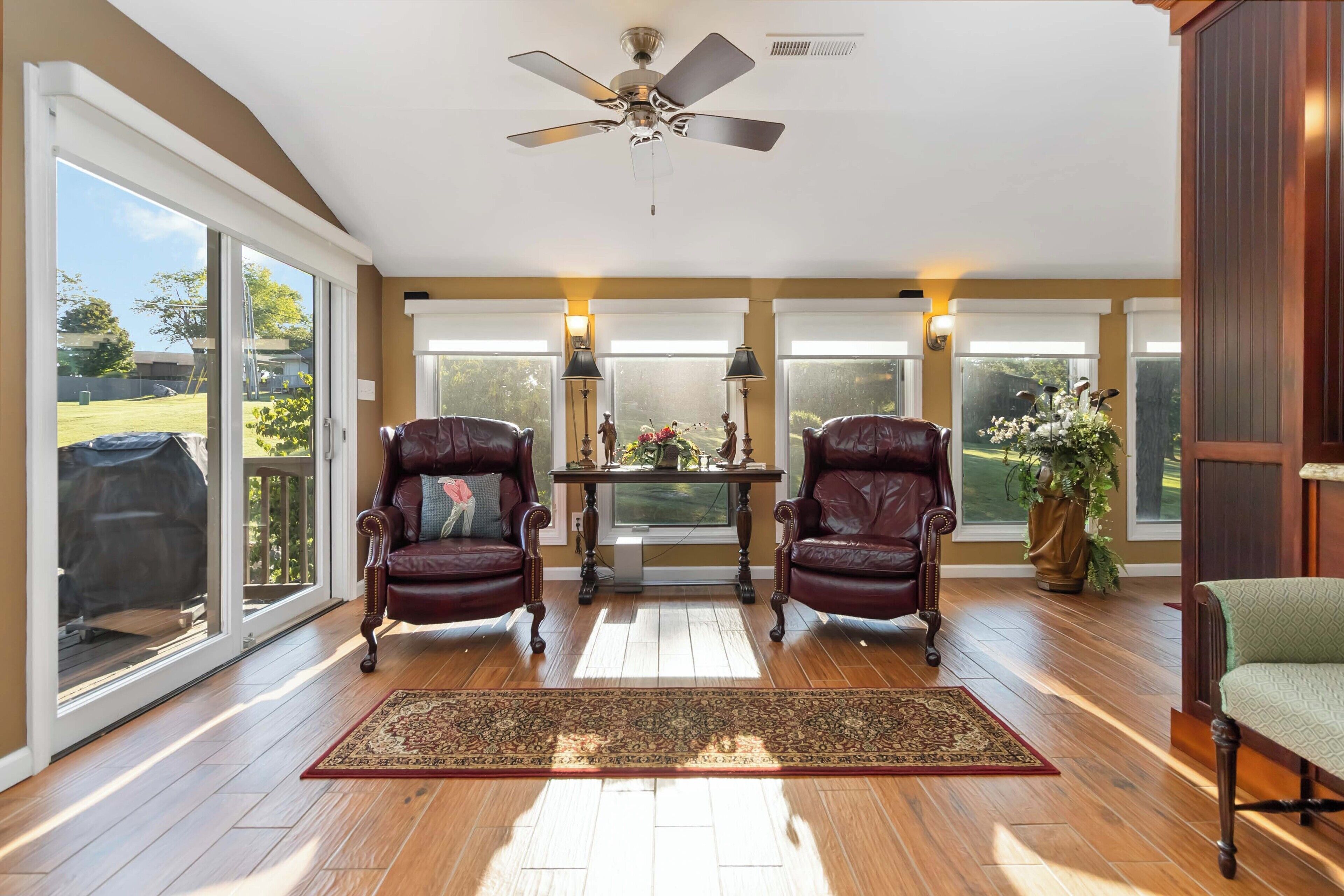 A spacious room with a ceiling fan, large windows, two leather chairs, a table with a vase, and a green chair.