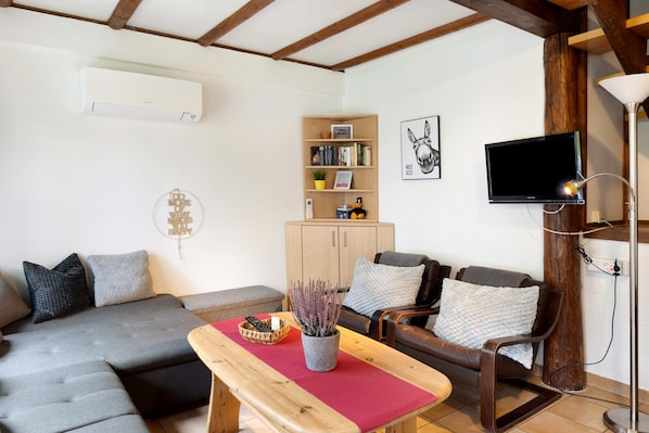 A living room with a sofa, a wooden table with a red runner, a bookshelf, a TV, and a lamp.
