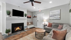 A living room with a fireplace, a ceiling fan, a grey sofa, and a painting on the wall. - 3BR Riverfront Suite w/ Full Kitchen & Game Room (Intervale)
