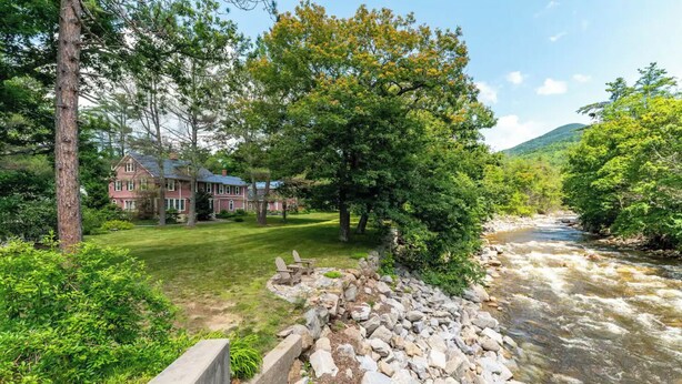 A riverside view with a house, a grassy area, and a rocky riverbank. - 3BR Riverfront Suite w/ Full Kitchen & Game Room (Intervale)