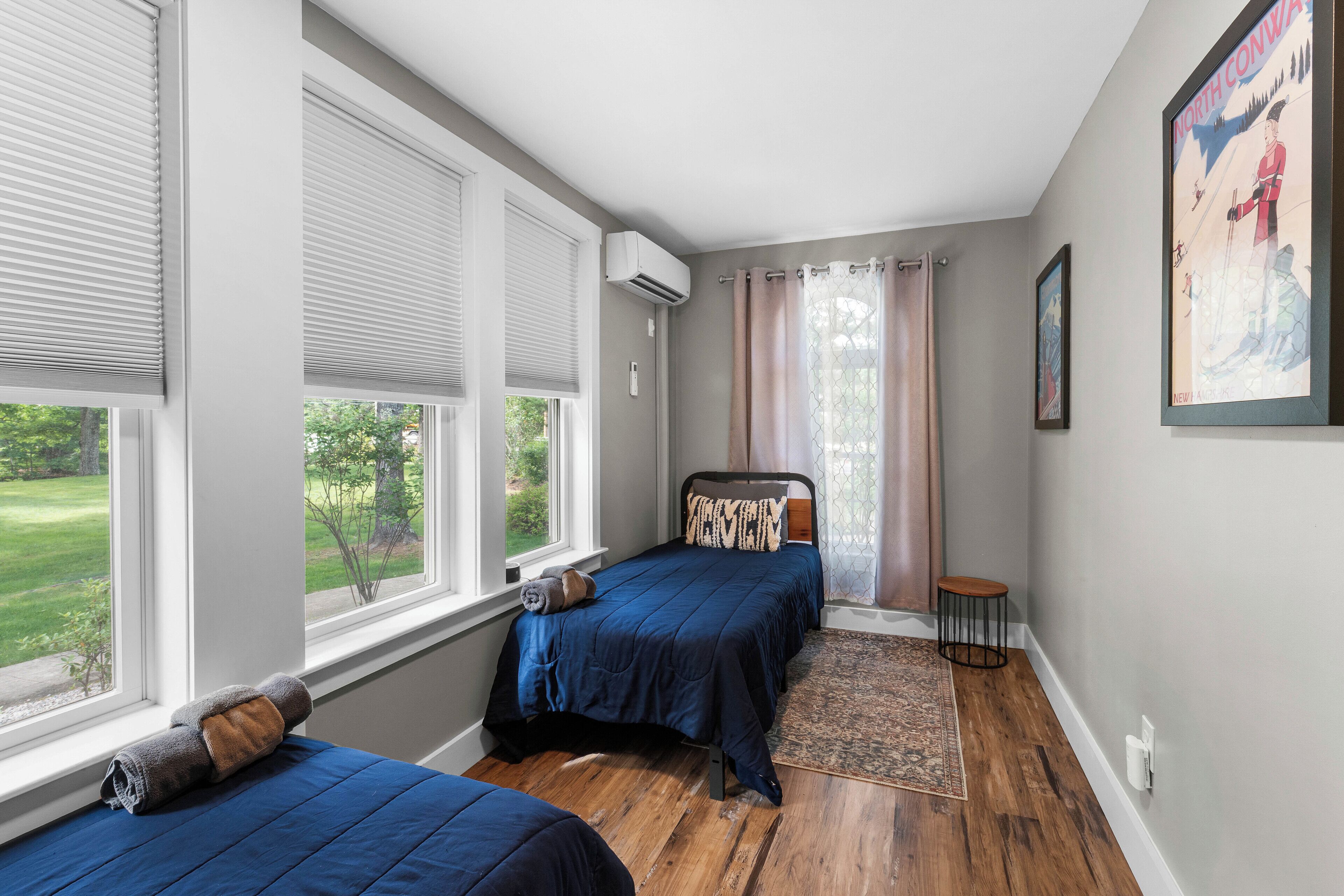 Une chambre avec deux lits, une fenêtre avec des stores, un petit tapis et un tableau encadré au mur.