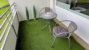 A balcony with artificial grass, a small table with chairs, and a window with a view of a green field.