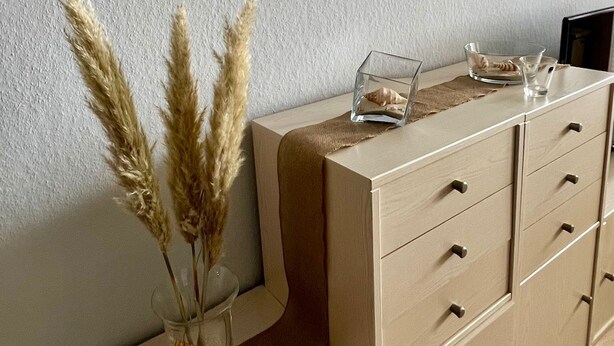 A room with a beige dresser, a vase with dried pampas grass, and two framed pictures on the wall.