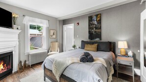 Een slaapkamer met een open haard, een bed met uitzicht, een nachtkastje met lamp en een schilderij aan de muur.