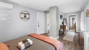 Une chambre avec un lit, une table de chevet, un climatiseur fixé au mur, une porte blanche et un couloir menant à une salle de bain.