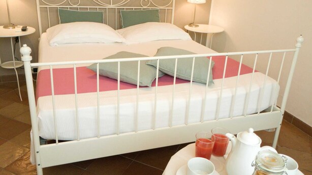 A bed with white metal headboard, two pillows, and a pink bedspread. There are two bedside tables with lamps, a tray with a teapot, cups, and a jar of sugar.