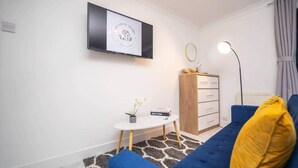 A modern living room with a blue sofa, a small white table with a plant, a wooden cabinet, and a flat-screen TV mounted on the wall. - Pleasant Studio in St Albans (St Albans)