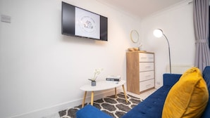 A modern living room with a blue sofa, a small white table, a wooden cabinet, and a flat-screen TV mounted on the wall.