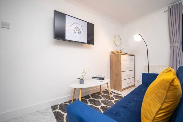 A modern living room with a blue sofa, a small white table, a wooden cabinet, and a flat-screen TV mounted on the wall.