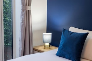 A hotel room with a bed, a nightstand with a lamp, a window with curtains, and a view of greenery outside.