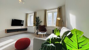 A modern living room with a flat-screen TV, a red knitted ottoman, a grey sofa, and a large green plant. - Favorite Stays - Düsseldorf (Düsseldorf)