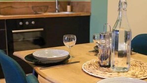 A dining table set with plates, cutlery, and glasses, with a bottle and pitcher on a woven placemat. A kitchen with a sink and stove is visible in the background. - A haven of peace near Pérouges, charm and comfort (Faramans)
