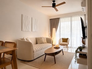 Un salon moderne comprenant un canapé, une table basse et des fauteuils, un ventilateur de plafond et une grande fenêtre avec des rideaux.