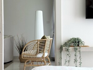 A room with a wicker chair, a tall white vase, and a plant on a shelf.