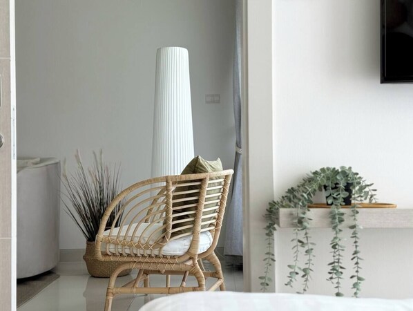 Una habitación con una silla de mimbre, un jarrón blanco alto y una planta en un estante.
