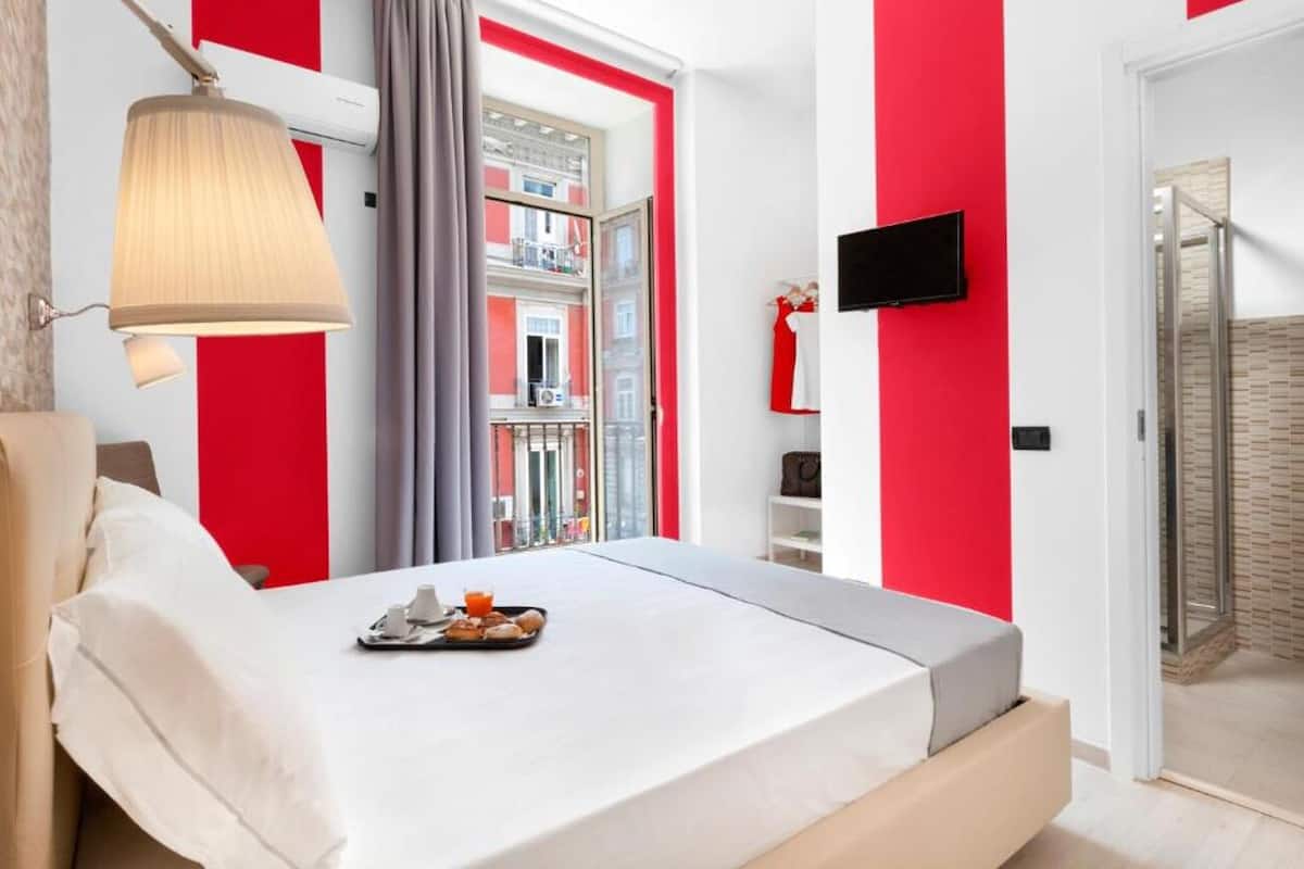 A hotel room with a bed, a bedside table with a tray of breakfast items, a TV mounted on the wall, and a view of the outside balcony.