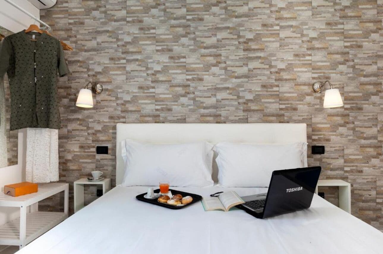 Ein ordentlich eingerichtetes Hotelzimmer mit Wandmuster, einem Bett mit weißer Bettwäsche, einem Laptop, einem Tablett mit Gebäck und Getränken sowie einem aufgehängten Kleidungsstück.
