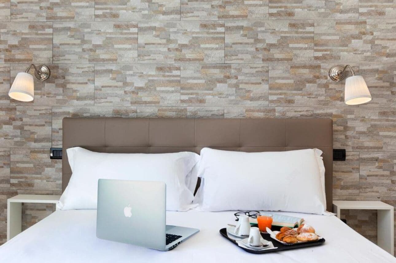 A hotel room with a stone-patterned wall, a bed with white linens, a laptop, and breakfast items on a tray.