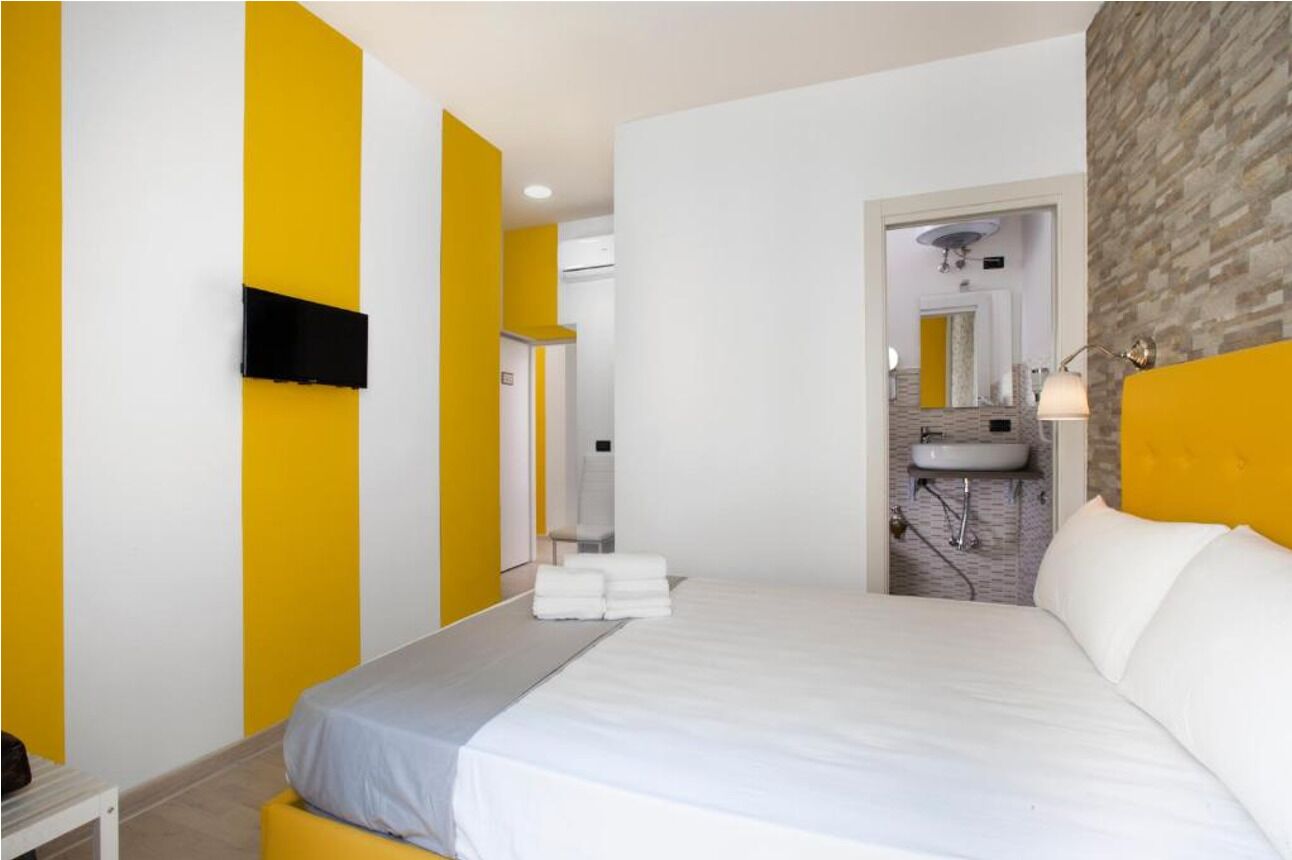 A modern hotel room with a bed, a flat-screen TV mounted on the wall, a bathroom with a sink and mirror, and a yellow accent wall.