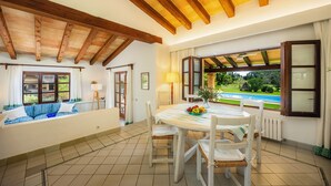 A dining area with a table and chairs, a window with curtains, and a view of a swimming pool. - Villa 'Reco Vert' with Mountain View, Private Pool and Air Conditioning (Pollença)