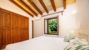 A bedroom with a large wooden wardrobe, a window with a view of greenery, a bed with white bedding, and a bedside lamp. - Villa 'Reco Vert' with Mountain View, Private Pool and Air Conditioning (Pollença)