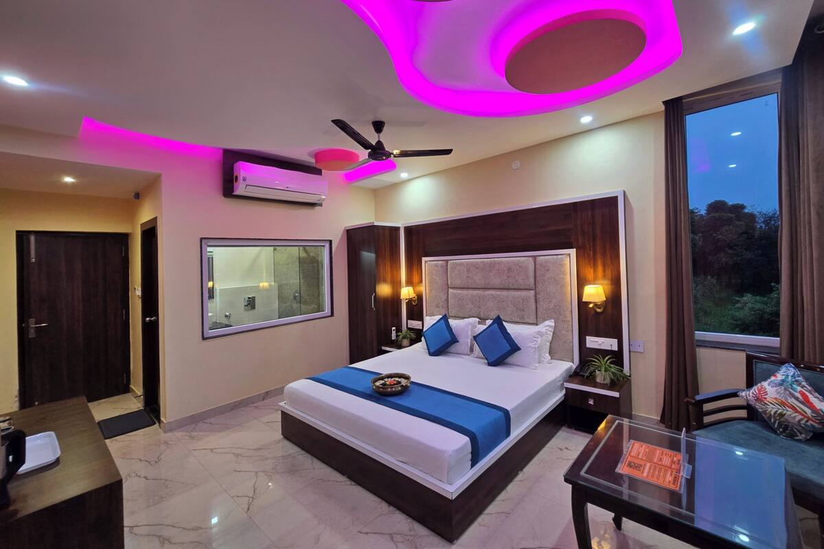 A modern bedroom with a large bed, a ceiling fan, an air conditioning unit, a window with a view, and a small table with a chair.