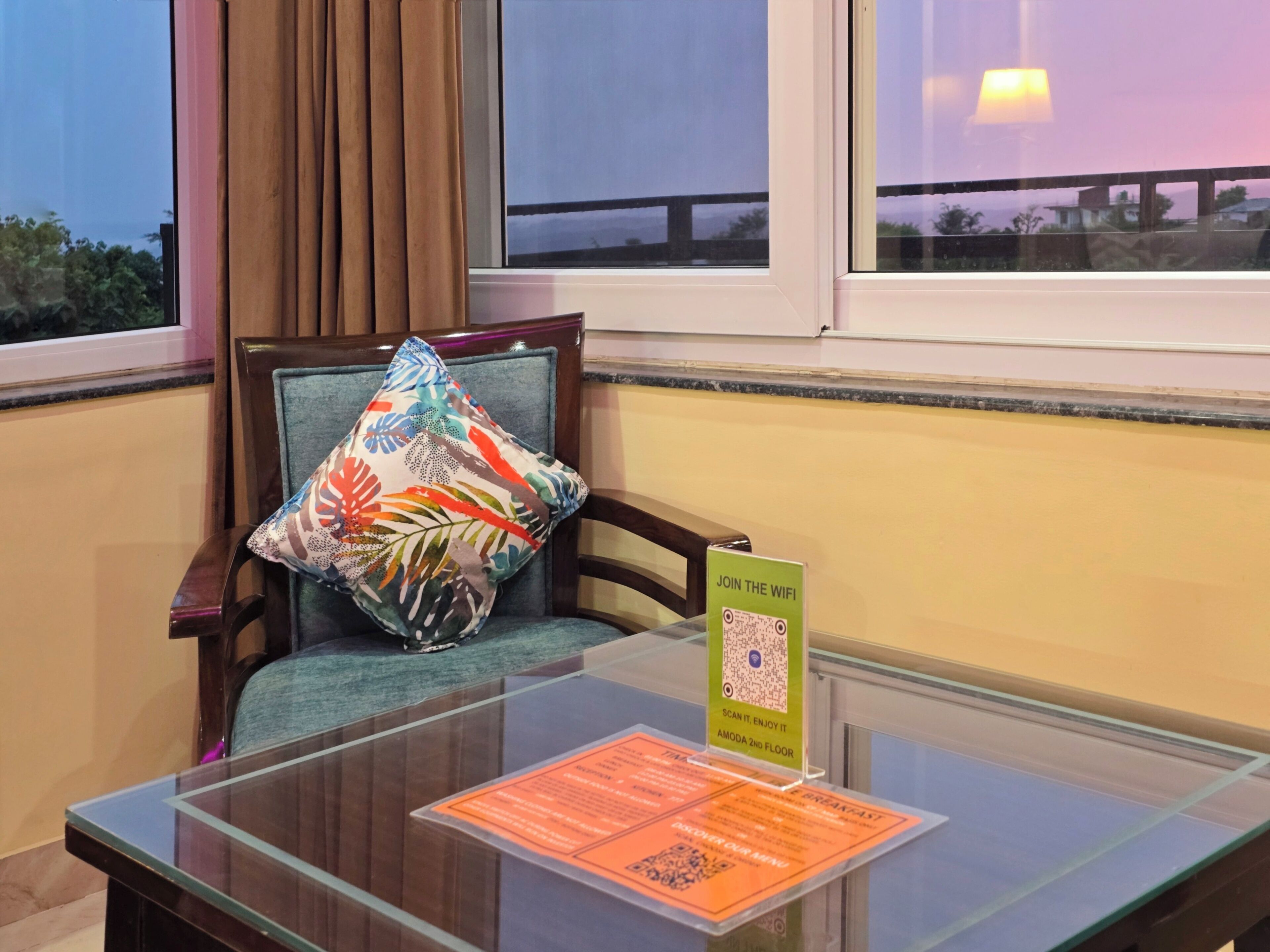 A hotel room with a glass table displaying a QR code for WiFi access, a colorful pillow on a chair, and a view of the outside through the window.