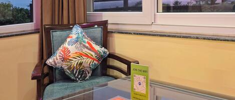 A hotel room with a glass table displaying a QR code for WiFi access, a colorful pillow on a chair, and a view of the outside through the window.