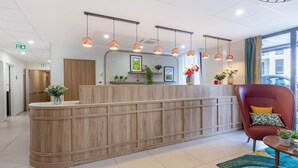 A reception area with a wooden reception desk, pendant lighting, a red armchair, and a view of the outside through large windows. - Zenao Appart'hôtels Tours (Tours)