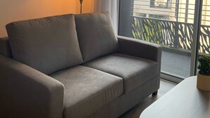 A grey sofa, a small table with a plant, a lamp, and a window with a view of buildings. - Zenao Appart'hôtels Tours (Tours)