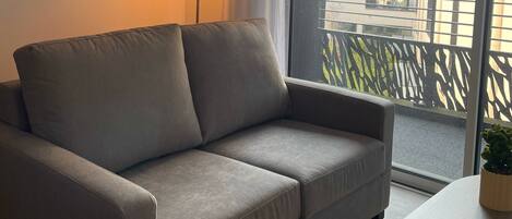 A grey sofa, a small table with a plant, a lamp, and a window with a view of buildings.