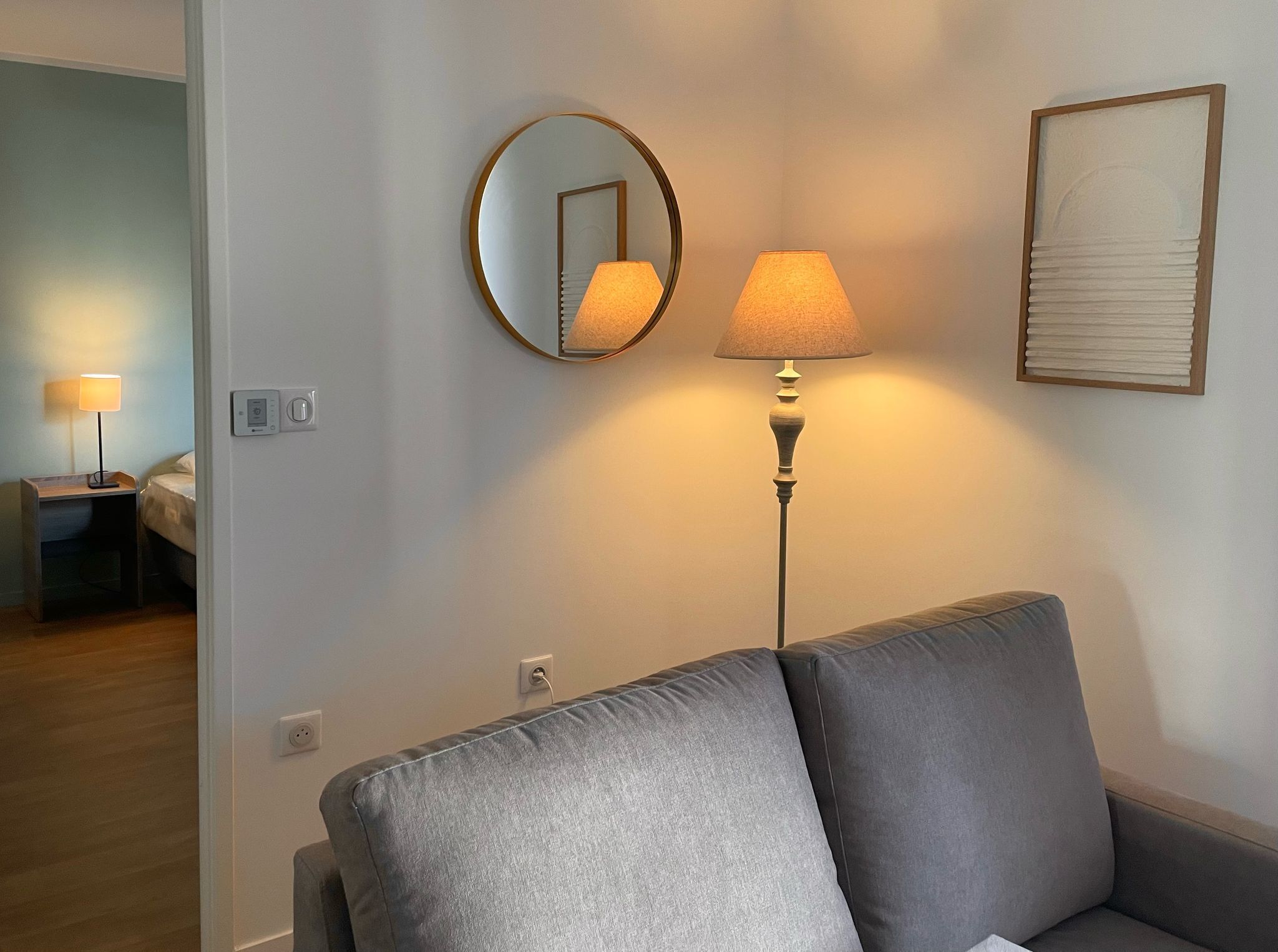 En moderne stue med en grå sofa, et rundt spejl, en gulvlampe og et indrammet kunstværk på væggen.