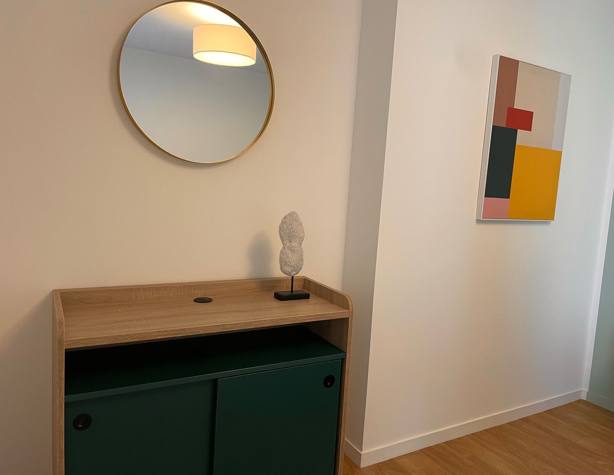 A room with a wooden console table, a green cabinet, a round mirror with a light, and a colorful abstract painting on the wall.