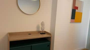 A room with a wooden console table, a green cabinet, a round mirror with a light, and a colorful abstract painting on the wall.