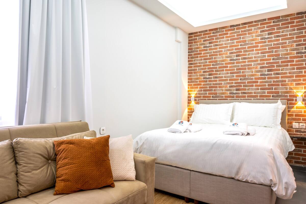 A hotel room with a bed, a sofa, a brick wall, and a skylight.