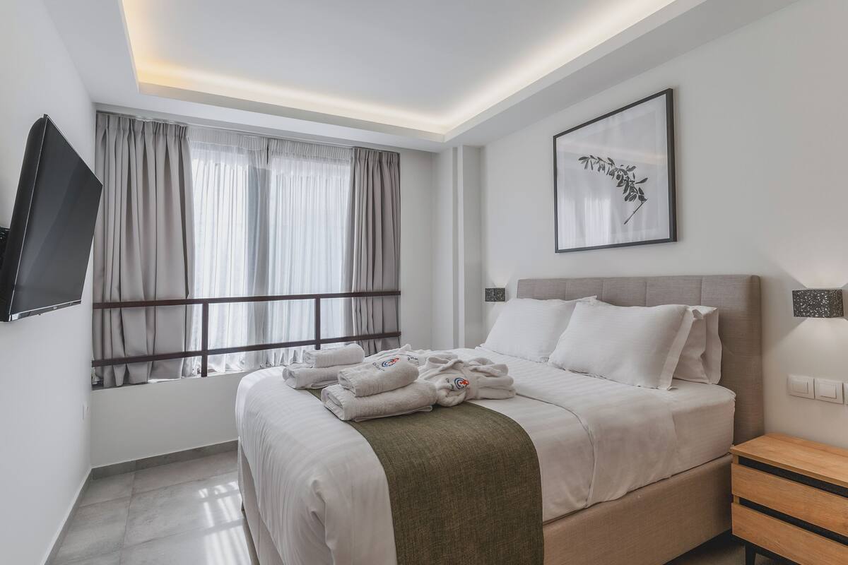 A hotel room with a bed, a TV mounted on the wall, a window with curtains, a framed picture, and a bedside table.