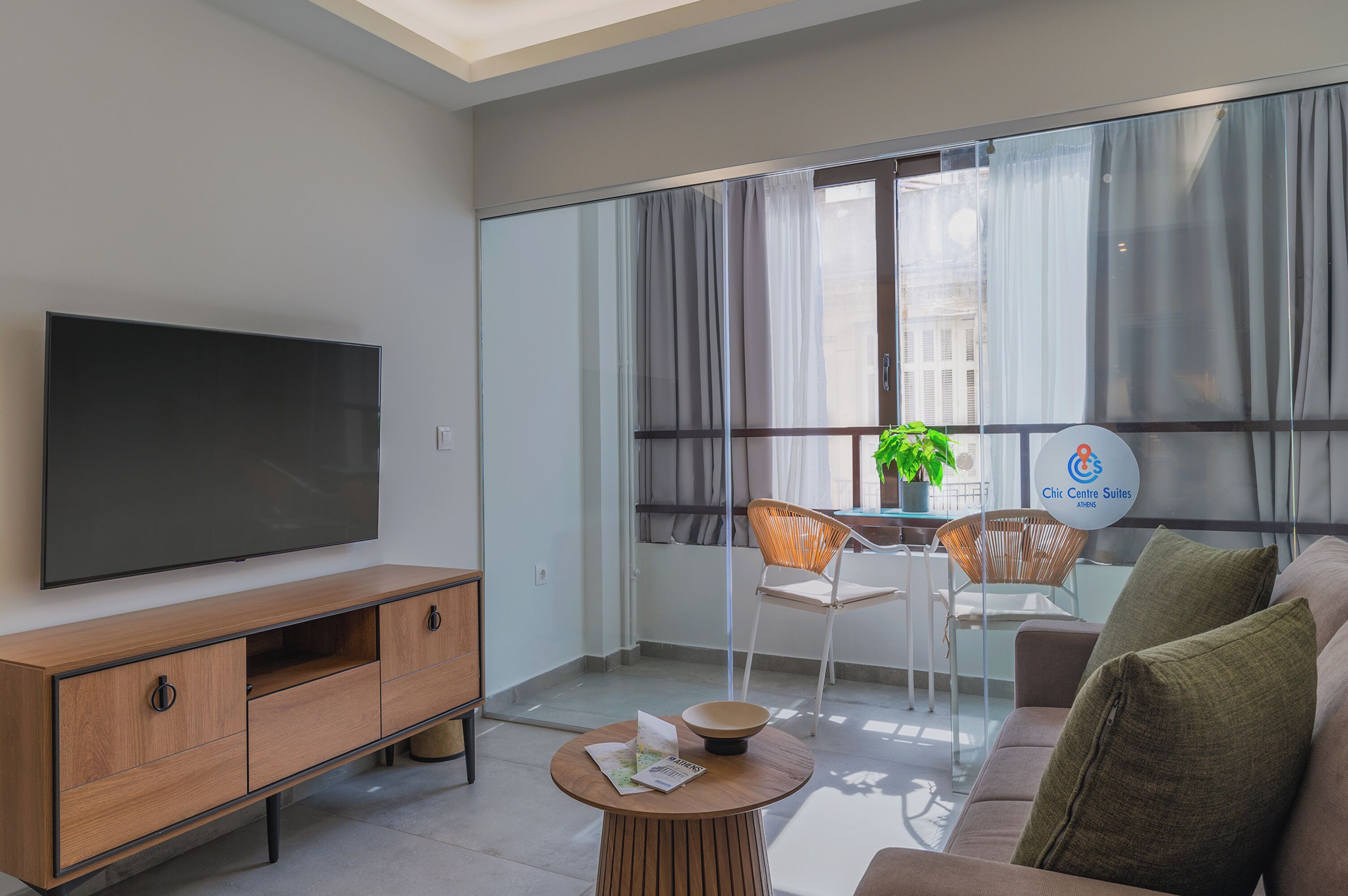 A modern living room with a flat-screen TV, wooden furniture, a round coffee table, and a view through large windows.