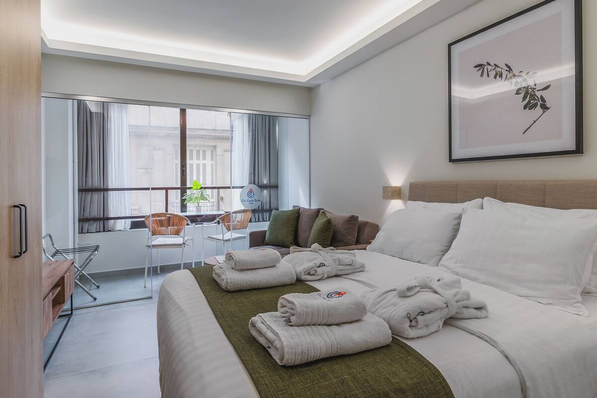 A hotel room with a bed, a chair, a desk, a window with curtains, and a framed picture on the wall.