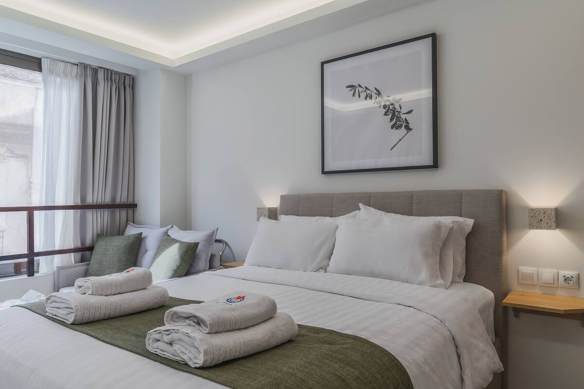 A hotel room with a bed, pillows, a bedside table, a wall-mounted light, and a framed picture.