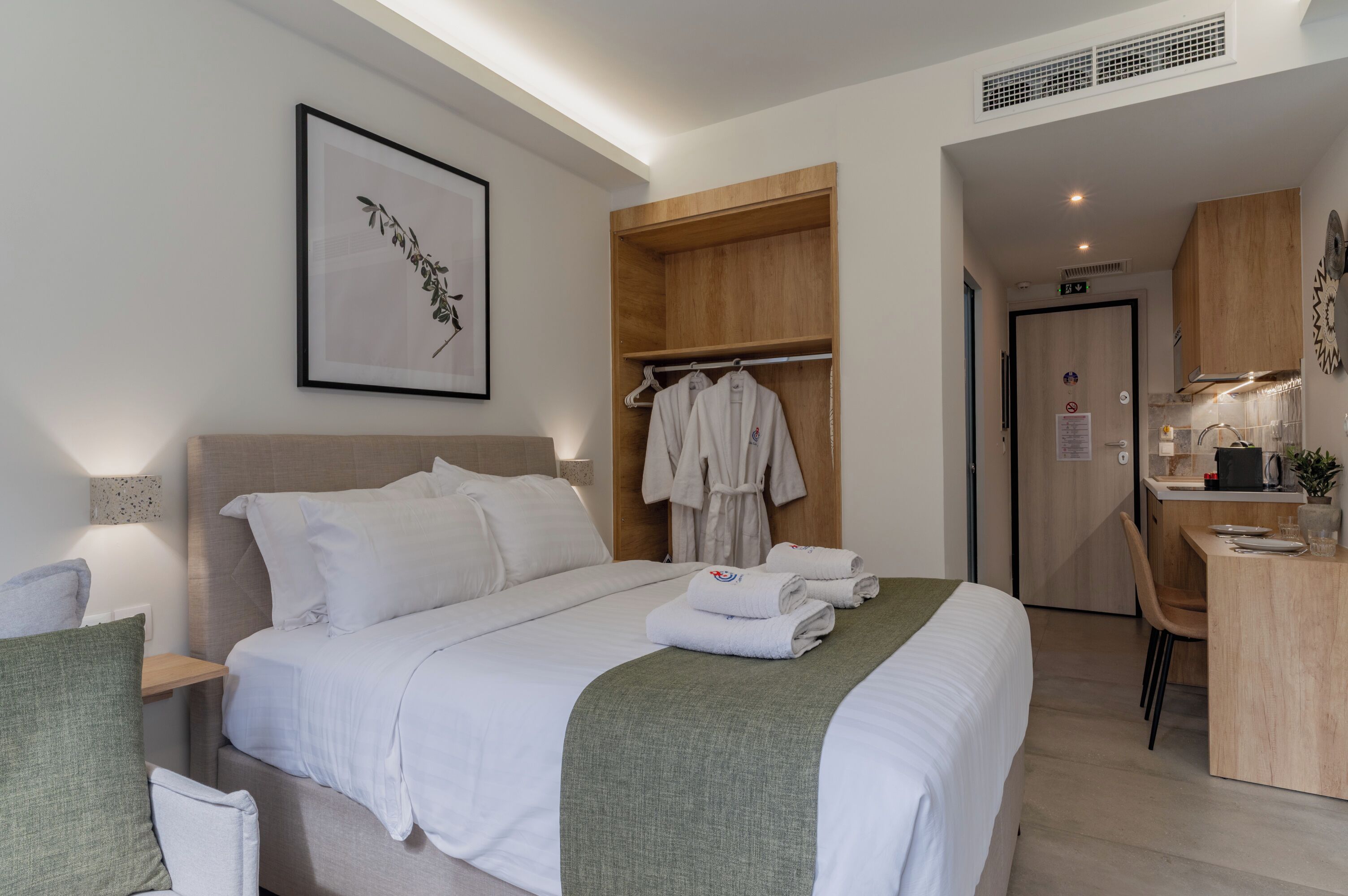A modern hotel room with a neatly made bed, a robe hanging on a wooden rack, a desk with a chair, and a framed artwork on the wall.