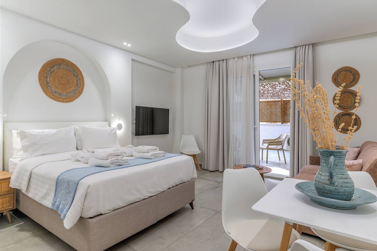 A modern hotel room with a bed, a flat-screen TV, a dining table, and a vase with dried plants.