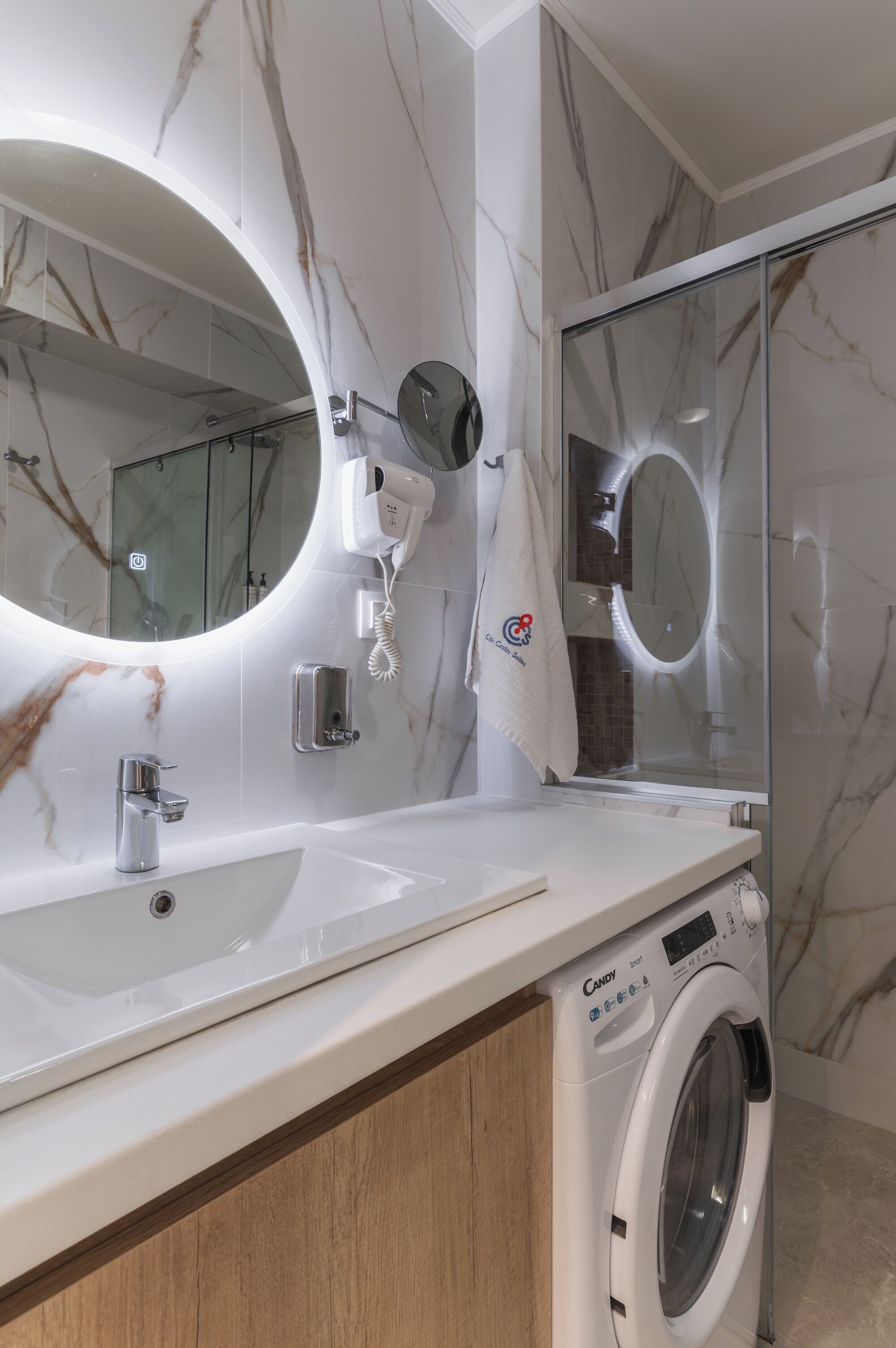Un bagno moderno con parete in marmo, uno specchio rotondo, un lavandino, un porta asciugamani, un asciugamani ad aria e una lavatrice.