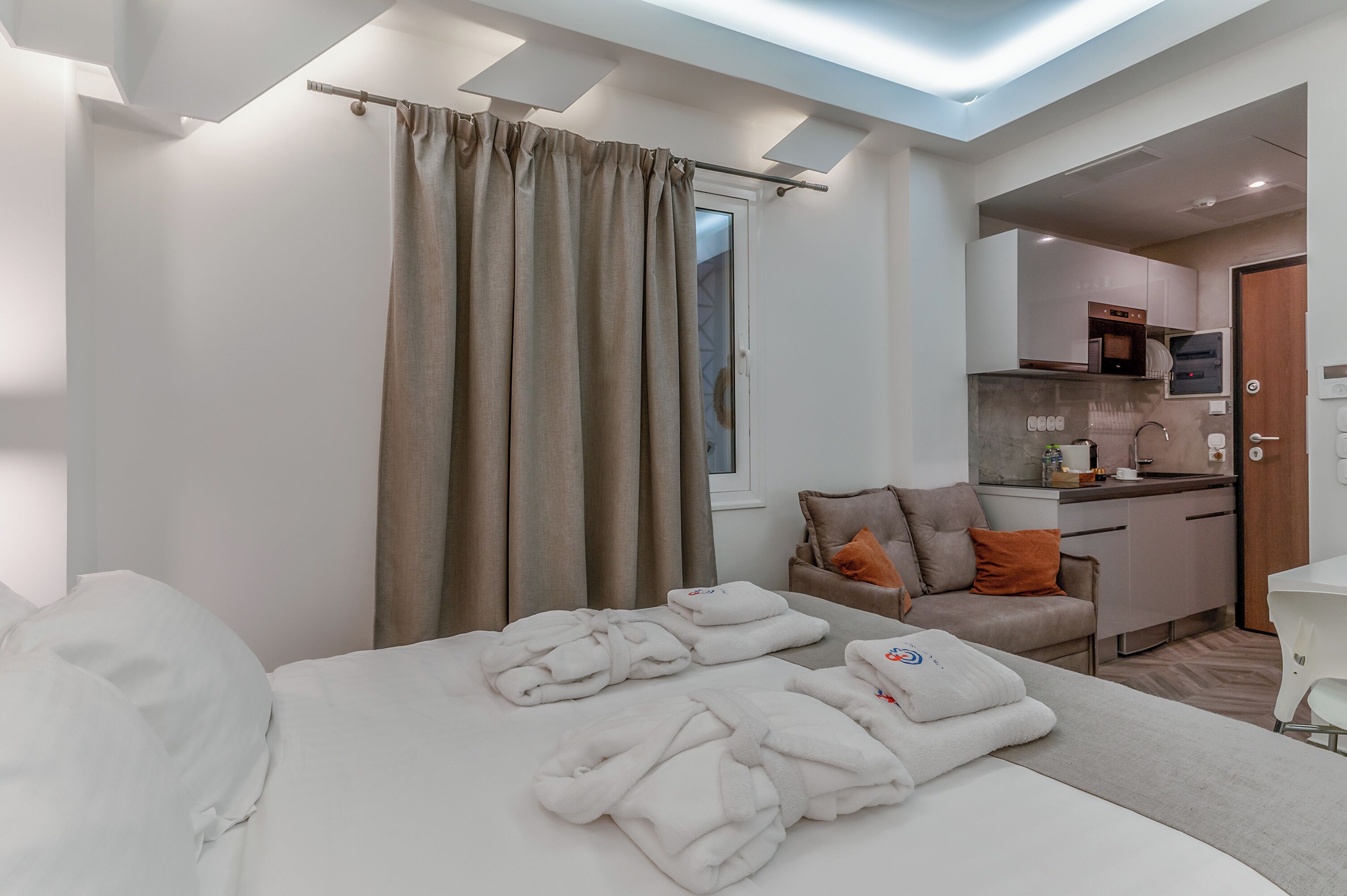 A hotel room with a bed, towels, a sofa, a kitchenette, and a window with curtains.