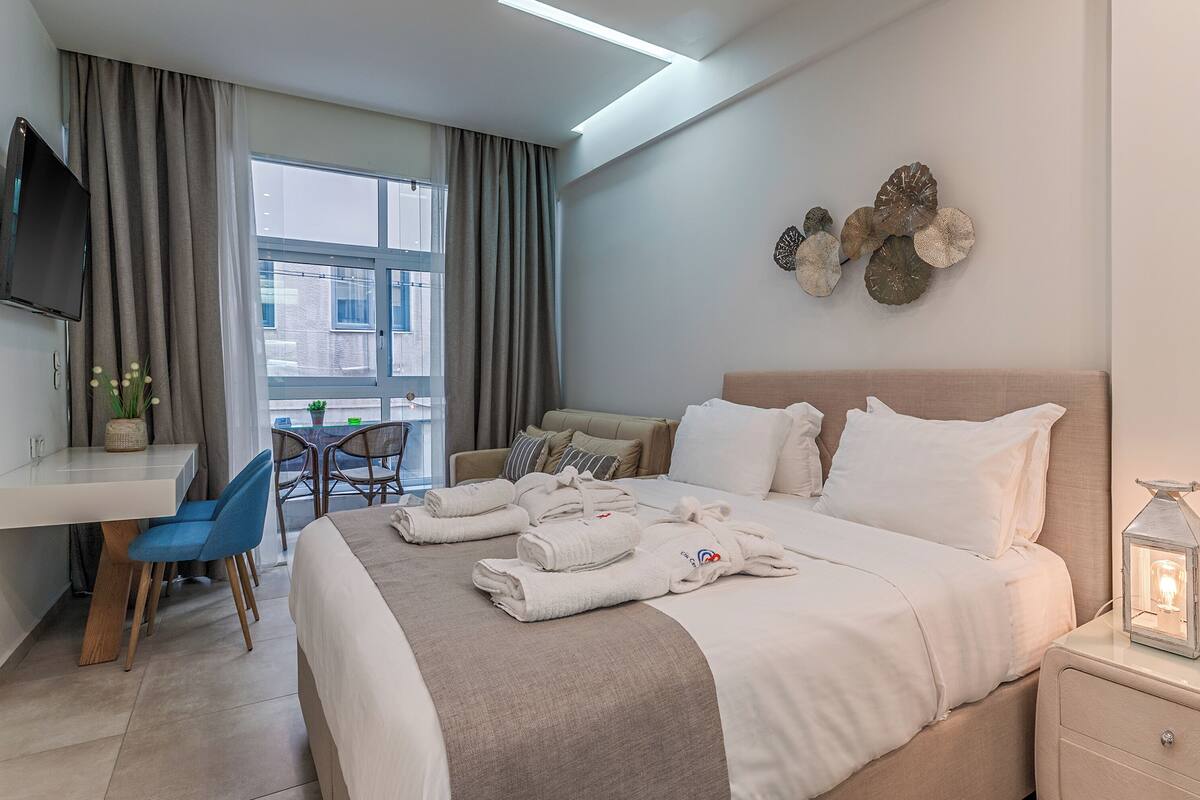 A hotel room with a bed, bedside table, TV, desk, chair, and a view of the city.