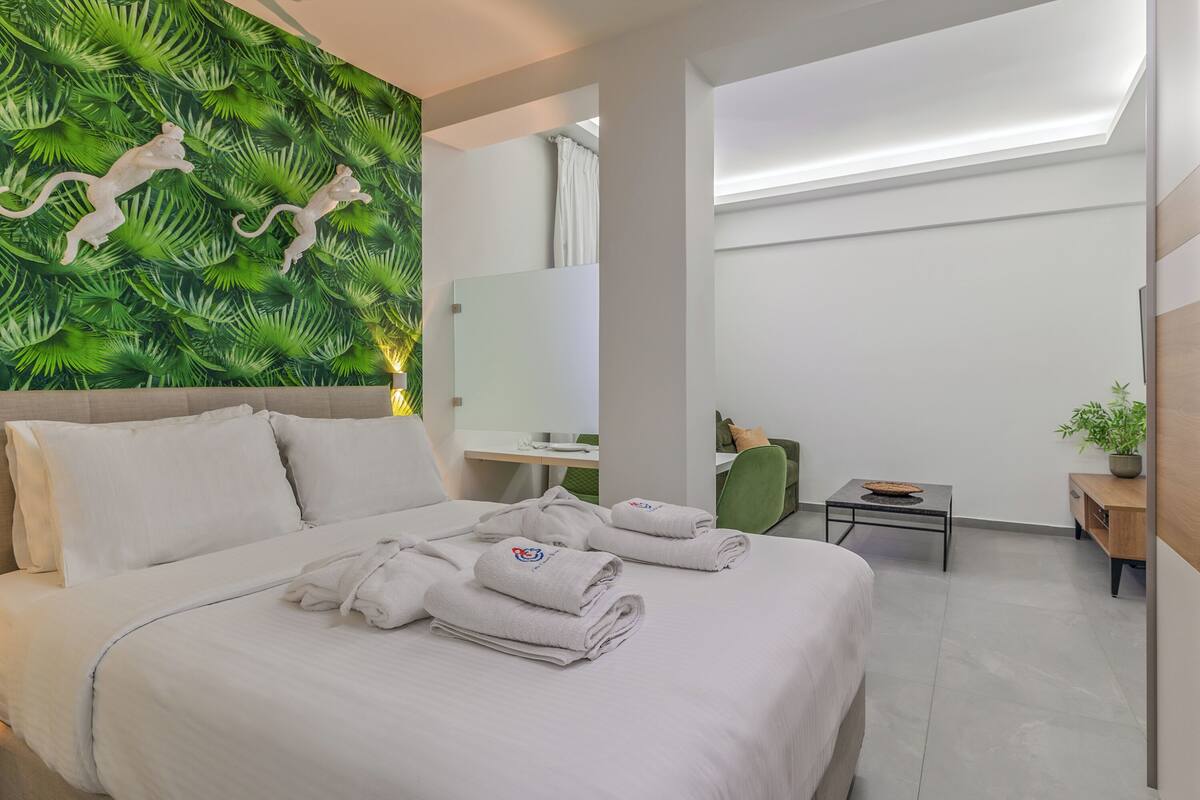 A hotel room with a bed, a green wall with animal decorations, a mirror, a desk, a chair, a small table, and a plant.