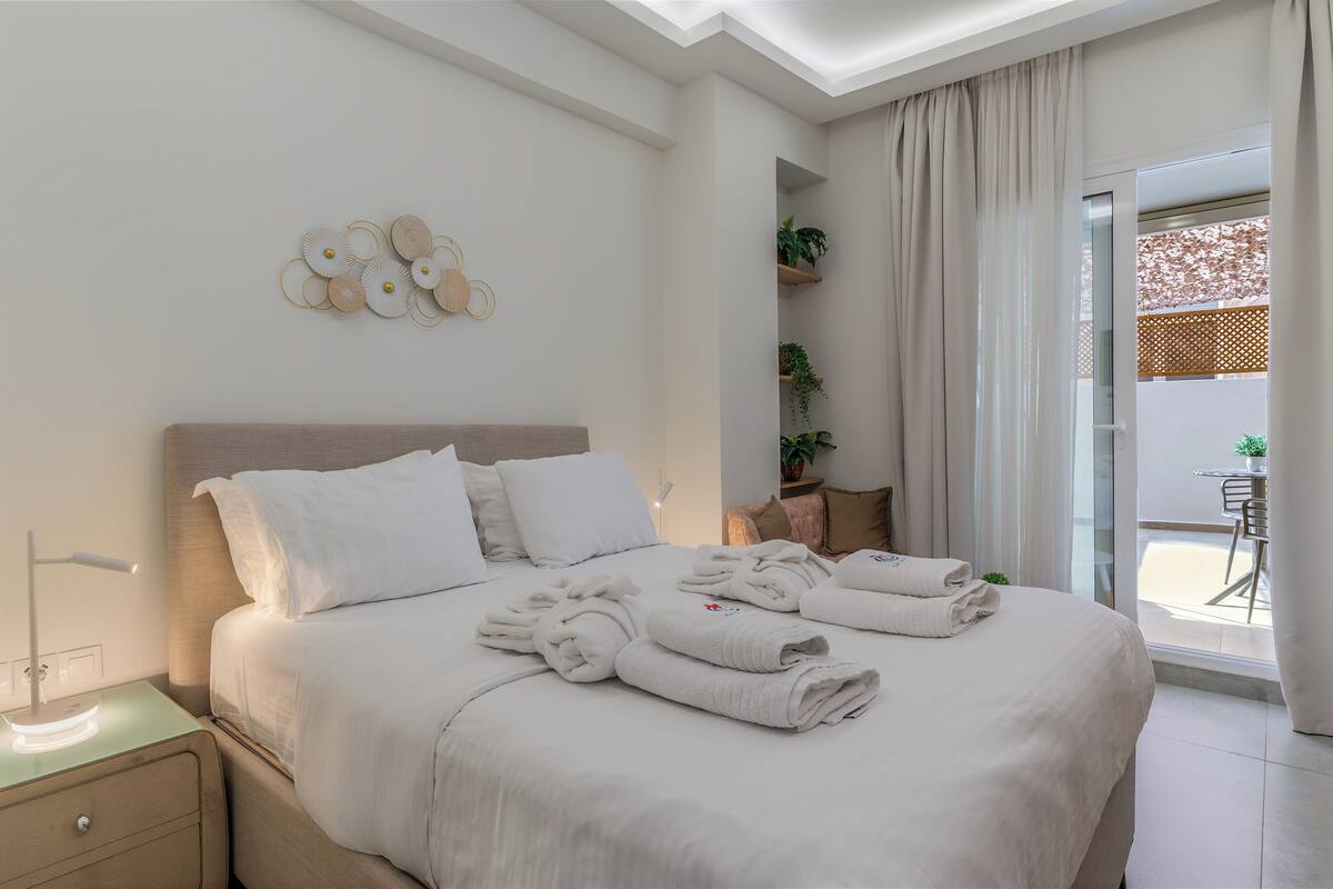 A hotel room with a neatly made bed, a bedside table with a lamp, a wall-mounted decorative piece, and a view of the outside through a large window.